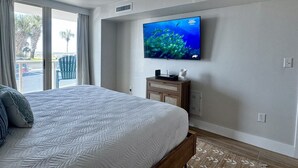 A hotel room with a large bed, a flat-screen TV mounted on the wall, a wooden nightstand, and a view of palm trees through the window. - Sabine 1E Couples Getaway Freshly Remodeled- Brand New Everything! (Pensacola Beach)