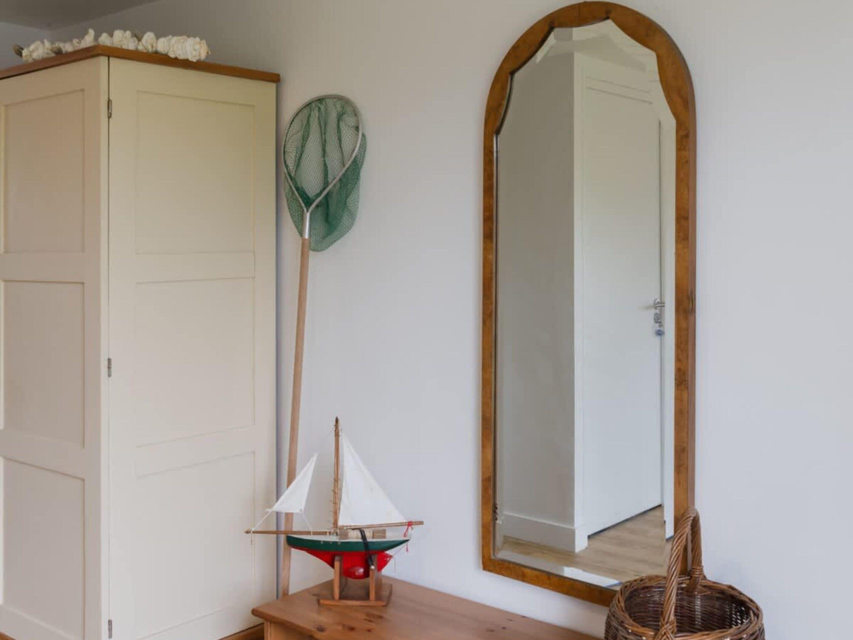A room with a white wardrobe, a green net, a wooden sailing ship model, a large mirror, and a wicker chair.