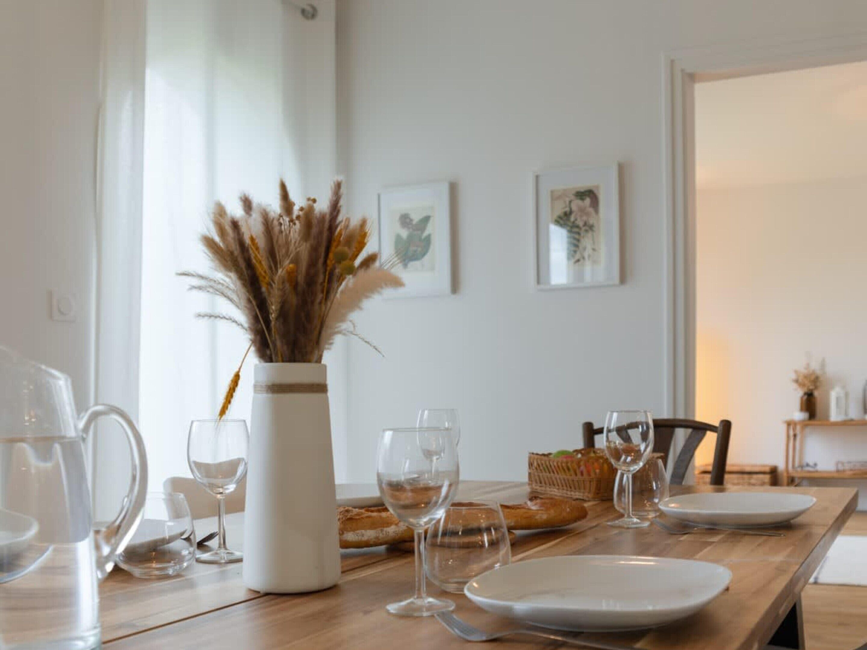 A dining table set with plates, glasses, and a pitcher, with a vase of dried plants on the table.