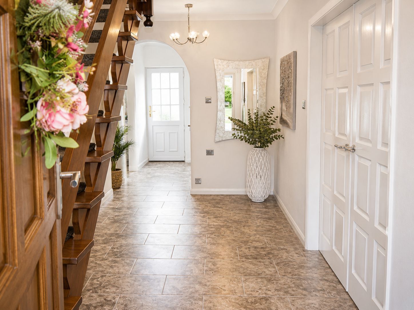 A hallway with a wooden staircase, a large white door, a mirror, and a potted plant.