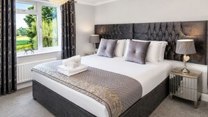 Una cama bien hecha con edredón gris y blanco, una lámpara de noche y vistas a zonas verdes al exterior.
