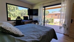 Une chambre à coucher comprenant un lit, un bureau avec un ordinateur, une télévision et une vue sur un jardin, accessible par une porte coulissante.