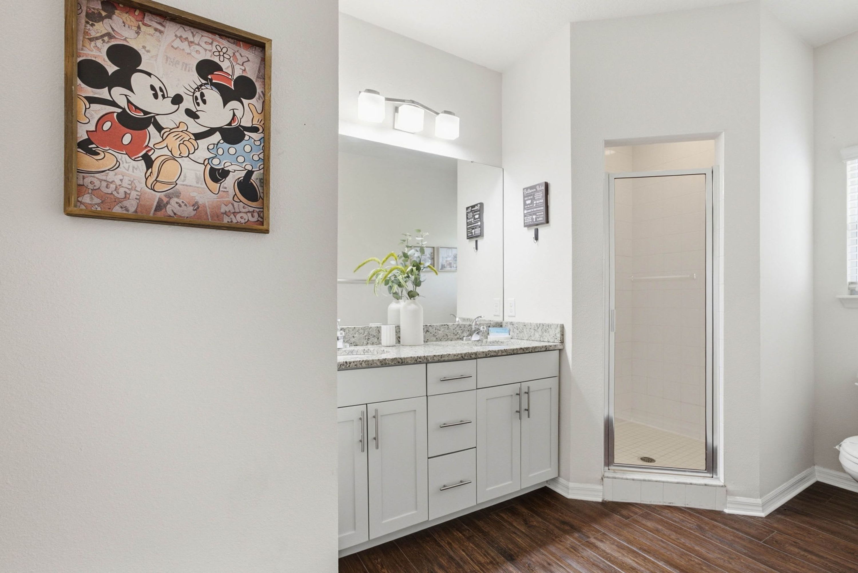 Une salle de bain décorée avec une œuvre d’art sur le thème de Mickey Mouse, un lavabo avec un miroir et un espace douche.