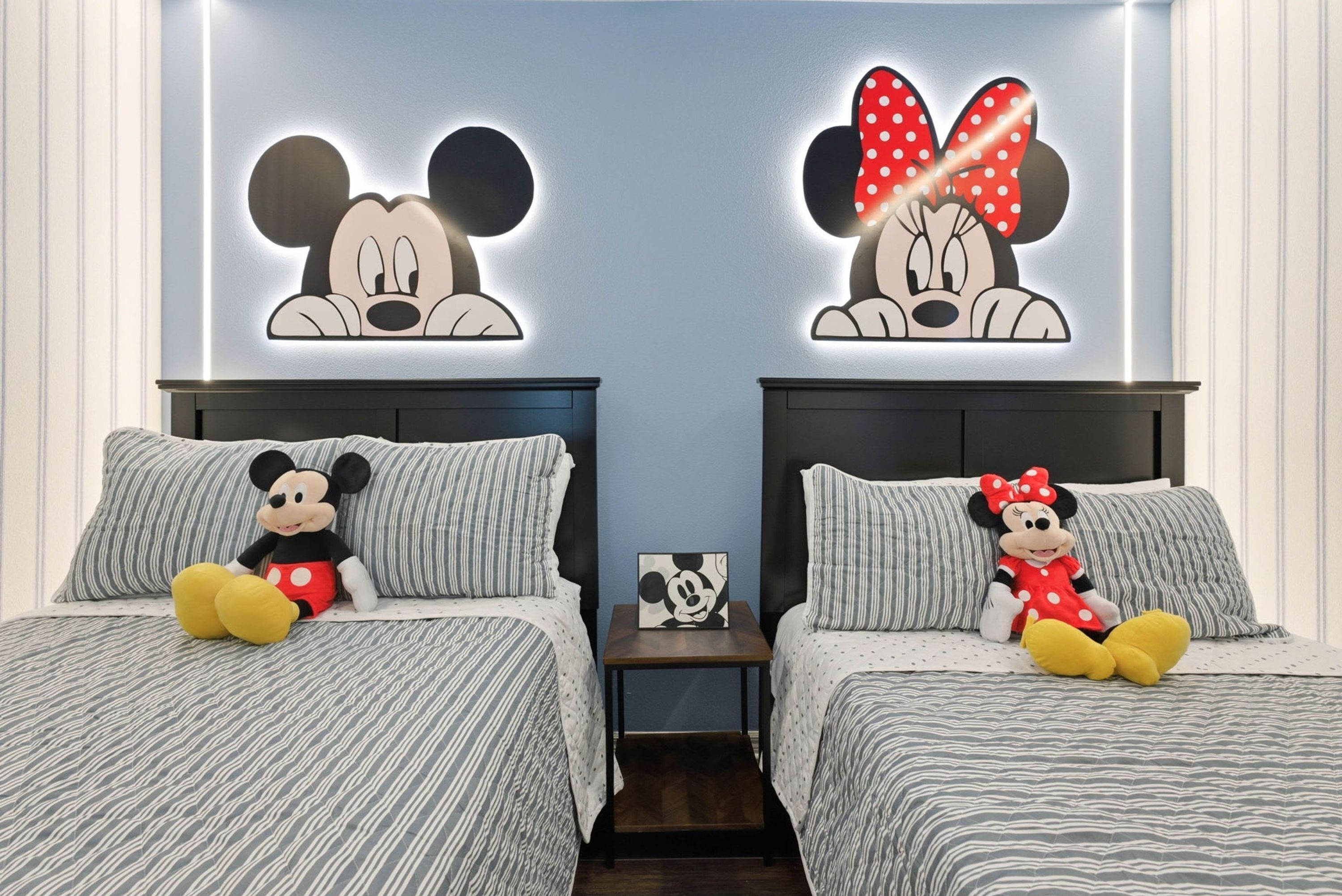 Two beds with Mickey Mouse and Minnie Mouse wall art, a nightstand with a Mickey Mouse picture, and striped bedding.