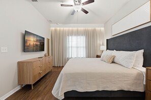 Ein Schlafzimmer mit einem Bett, einem Deckenventilator, einem Fernseher, einer Kommode und einem Fenster mit Vorhängen.