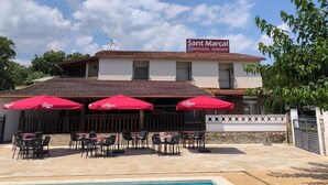 Ein Gebäude mit einem Schild, auf dem 'Sant Marçal HABITACIONS - RESTAURANT' steht, Außenbereich mit roten Sonnenschirmen und einem Schwimmbecken.