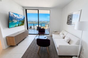 A modern living room with a sofa, a TV, a dining table, and a view of the sea through large windows. - Corner Bay-View Stay with Pool and Balcony in Miami G2515 (Miami Beach)