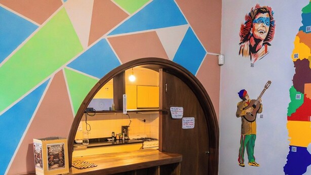Un coin repas avec une table et des chaises en bois, une cuisine visible à travers une arche, et des œuvres d’art colorées sur les murs, dont une représentation d’une personne jouant de la guitare.