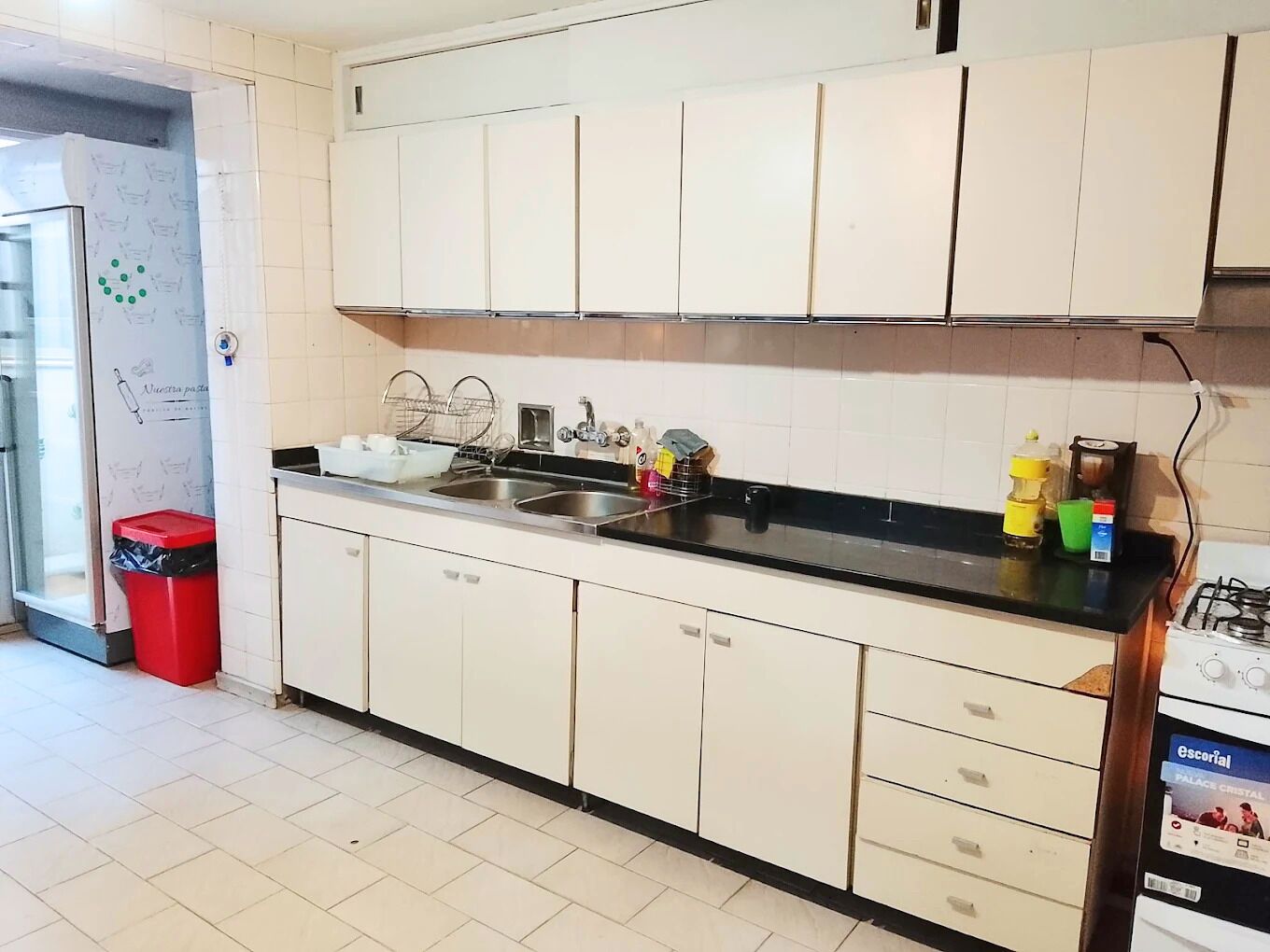 Una cocina con armarios blancos, encimera negra, pileta de acero inoxidable y un bote de basura rojo.
