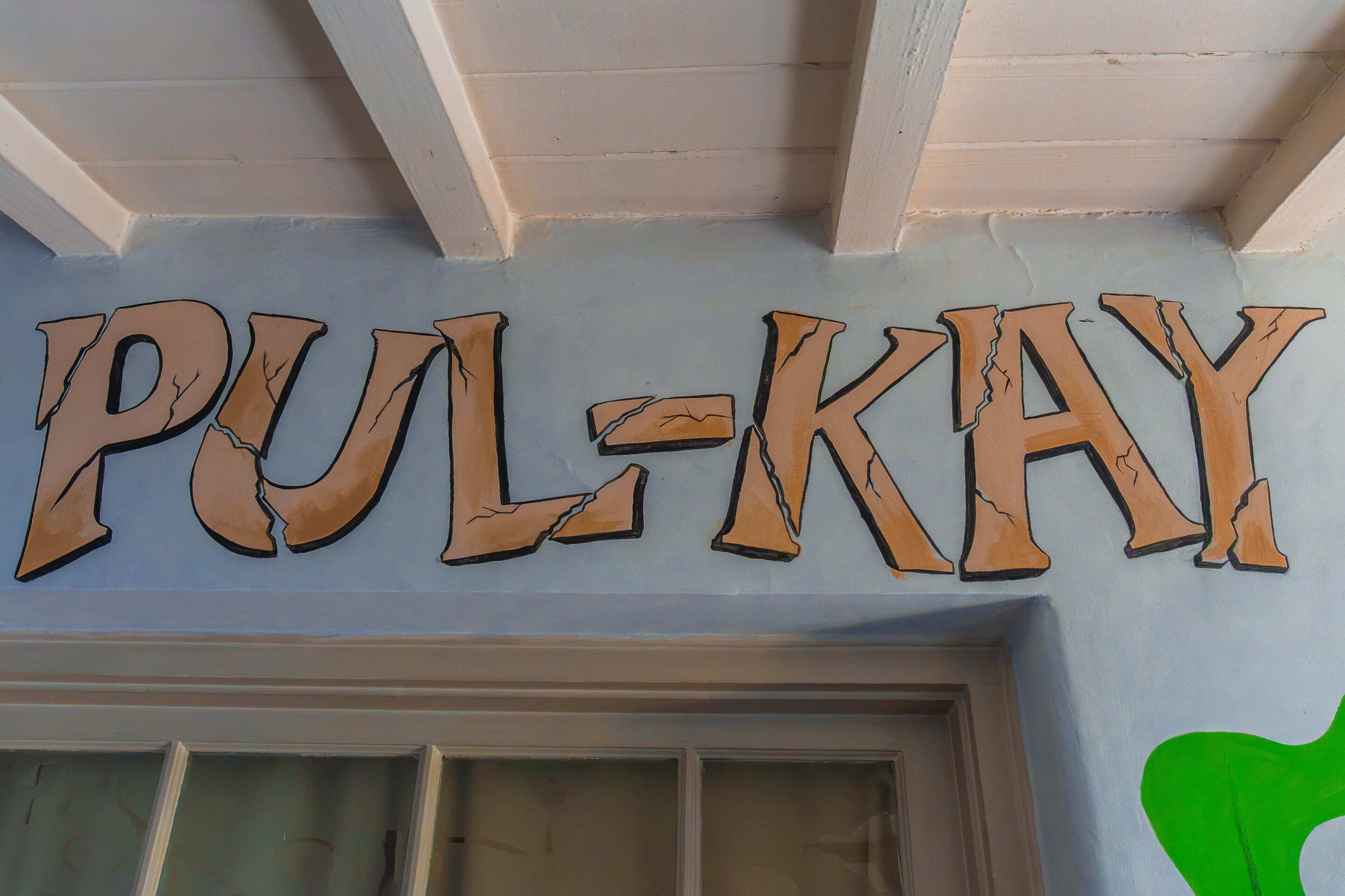 A sign with the text 'PUL-KAY' on a light blue wall.