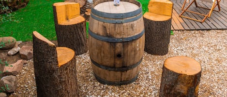 Un área de estar al aire libre de estilo rústico, con una mesa de barril de madera rodeada de taburetes, sobre un suelo de grava. Se aprecia un árbol y sillas al fondo.