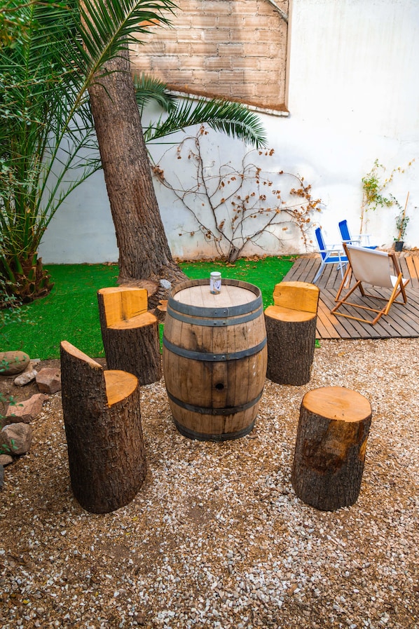 Un área de estar al aire libre de estilo rústico, con una mesa de barril de madera rodeada de taburetes, sobre un suelo de grava. Se aprecia un árbol y sillas al fondo.