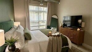 Ett sovrum med en säng, ett nattduksbord, en lampa, en tv, en byrå och ett fönster med persienner.