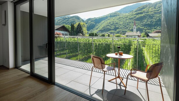 Un balcon avec une table et des chaises, une vue sur une vallée verdoyante et une église avec un clocher rouge.