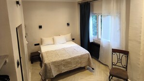 Un dormitorio con una cama, una silla, una mesita de noche, un ventanal con cortinas y un radiador.