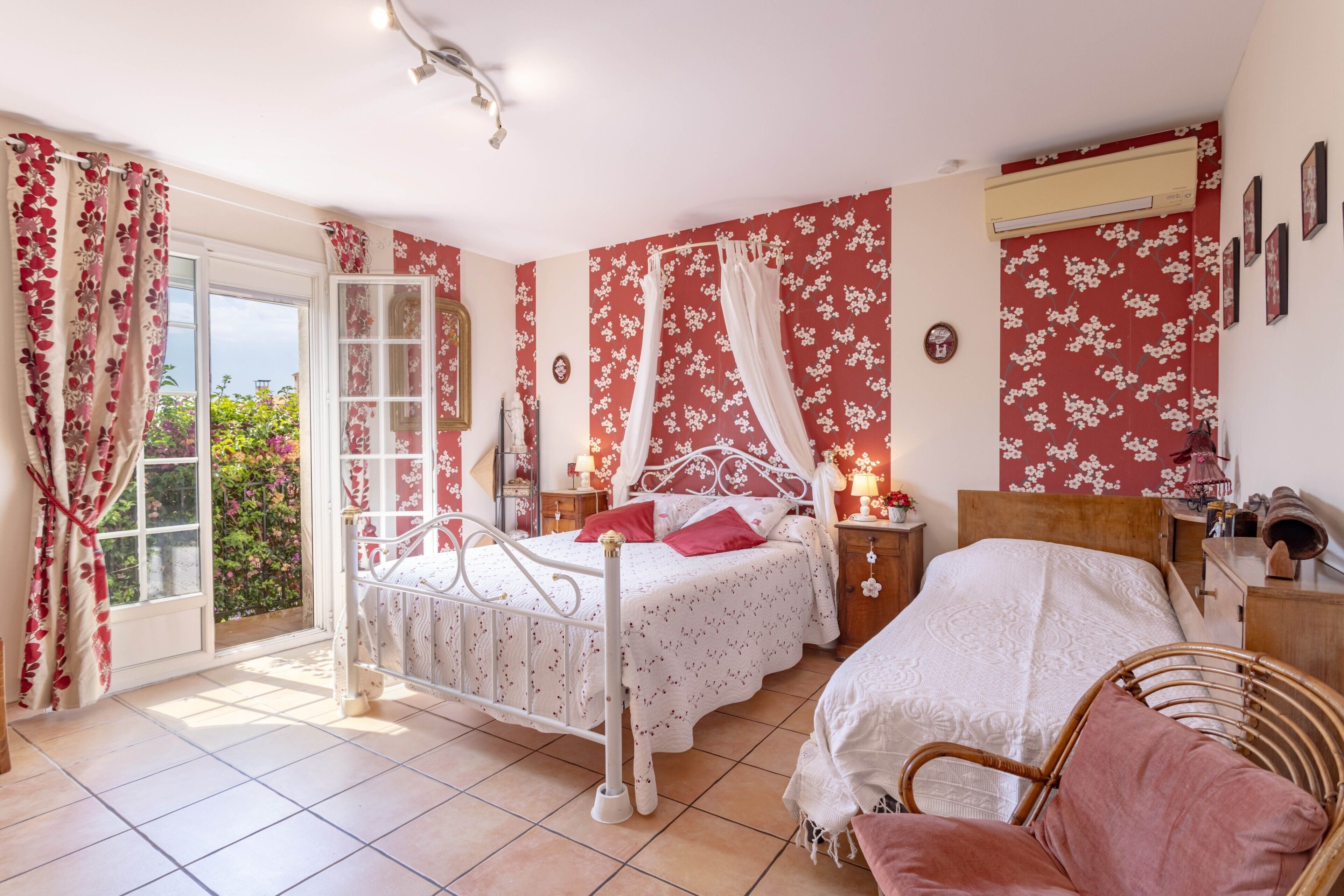 Een slaapkamer met een bed, een stoel, een nachtkastje, een raam met gordijnen en een deur die uitkomt op een balkon.