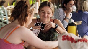 Eine Gruppe von Menschen spielt Karten an einem Tisch im Freien, im Hintergrund ein Holzzaun und viel Grün.