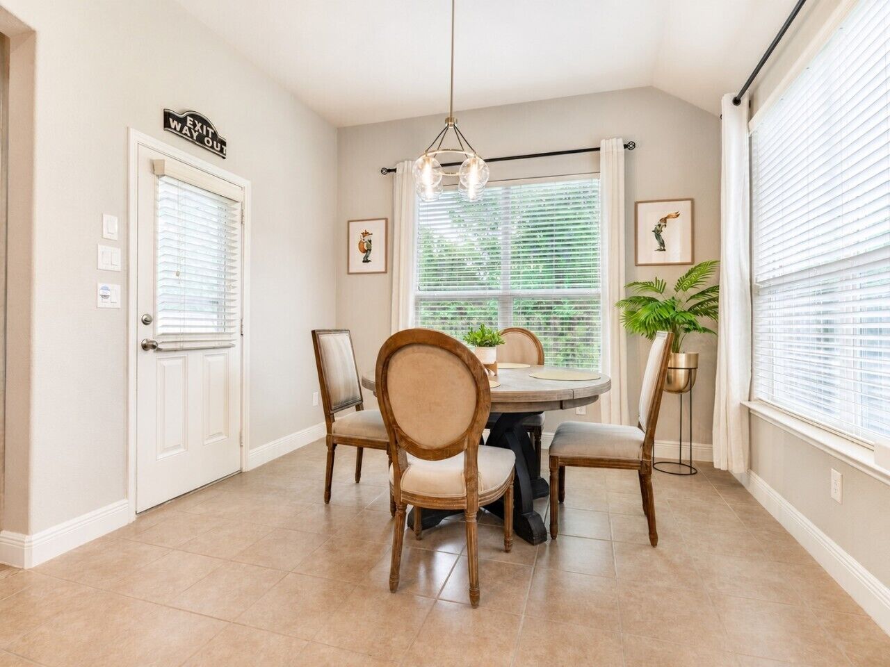 A dining area with a round table, four chairs, a hanging light fixture, a potted plant, and a window with blinds.