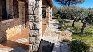 A stone building with a wooden porch, a view of olive trees, and a clear sky.