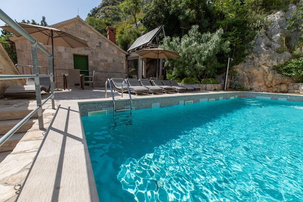 Ein Schwimmbecken mit Liegestühlen, ein Steinhaus mit grüner Tür und eine felsige Hügelkulisse im Hintergrund.