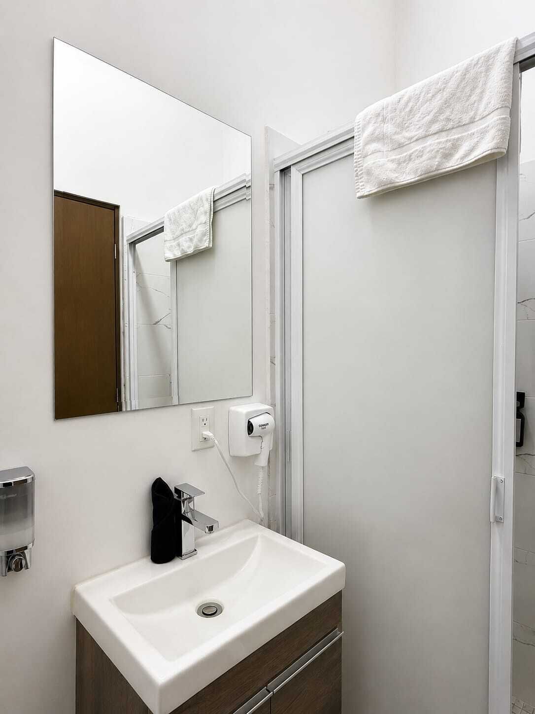 Un baño con un lavamanos blanco, un espejo, una toalla colgada en la puerta y un secamanos en la pared.