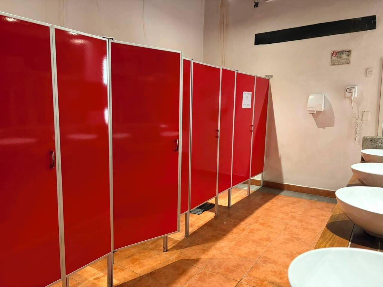 A row of red privacy cubicles in a bathroom with white sinks and a hand dryer.