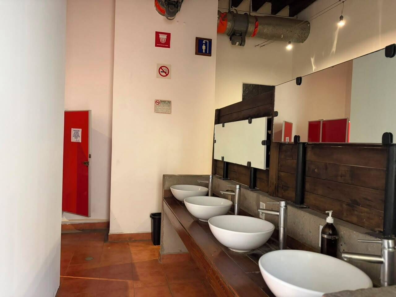 Eine öffentliche Toilette mit vier Waschbecken, einer roten Tür und einem Spiegel mit Holzrahmen.