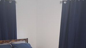 Ein Bett mit einer beigen Steudecke, zwei blauen Kissen, einem Holz-Kopfteil, einem kleinen Nachttisch und dunklen Vorhängen.