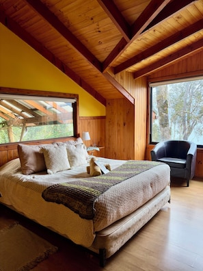 A bedroom with a large bed, a chair, and a window with a view of trees. - Turismo La Rueda del Chucao (Valdivia)
