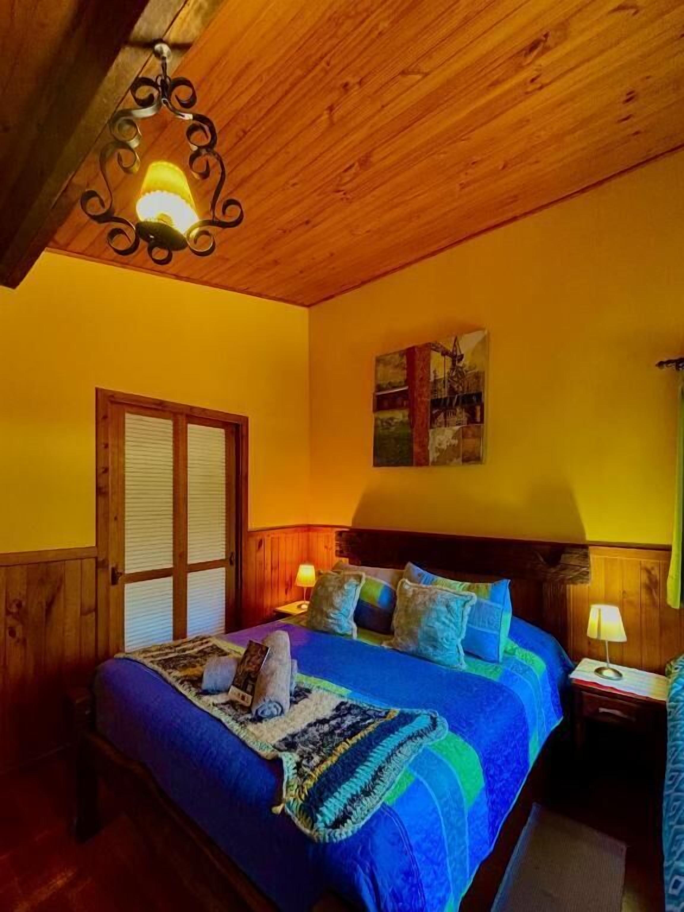 Una habitación con techo de madera, una cama con ropa de cama azul, una mesita de noche con lámpara y una ventana con contraventanas.