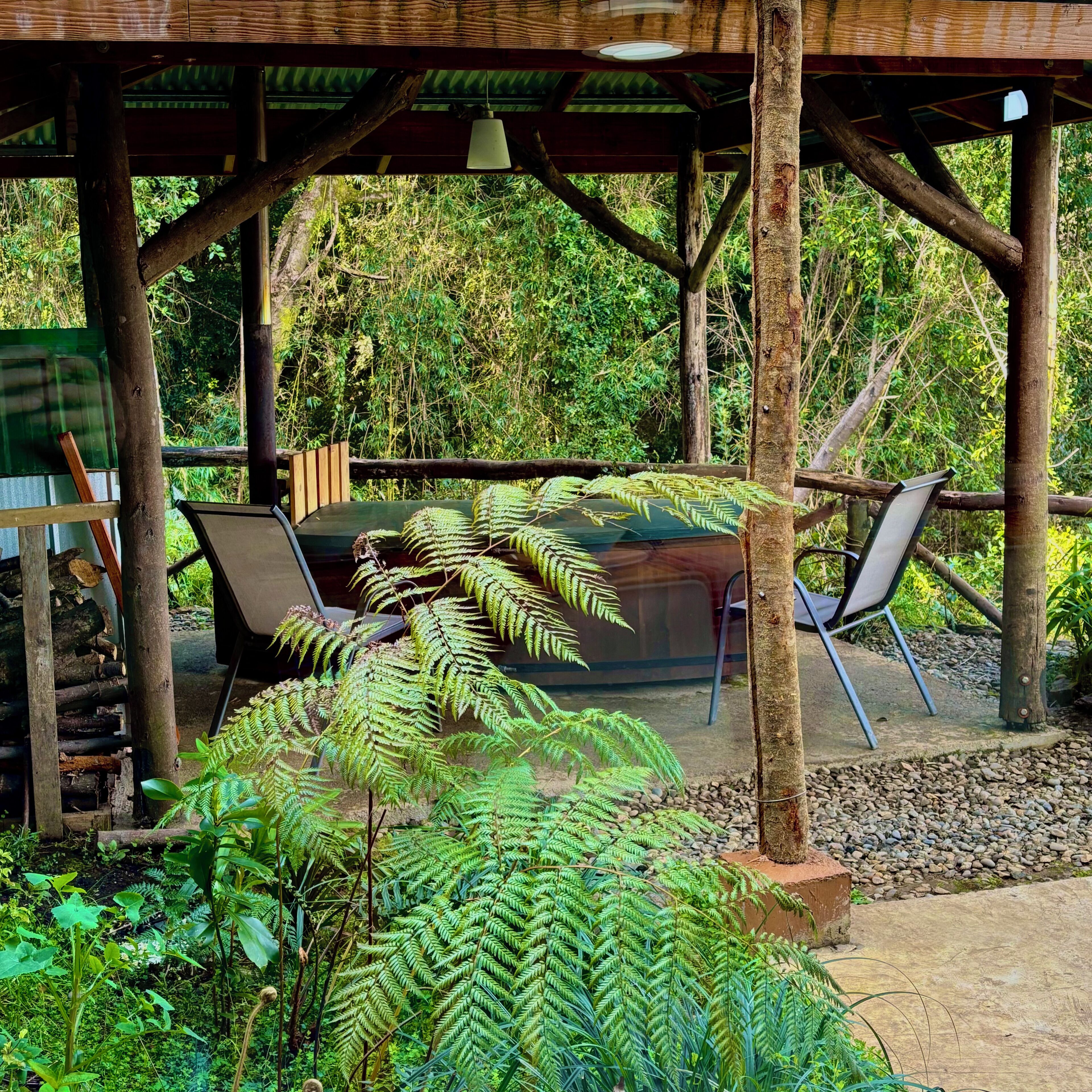 Ein hölzernes Pavillon mit Tisch und Stühlen, umgeben von üppigem Grün und einem Farn.