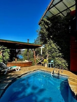 A swimming pool with a wooden deck, a covered seating area, and a view of trees and a building.