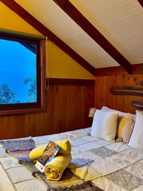 Un dormitorio con un gran ventanal, paredes de madera, una cama con colcha y una revista enrollada.