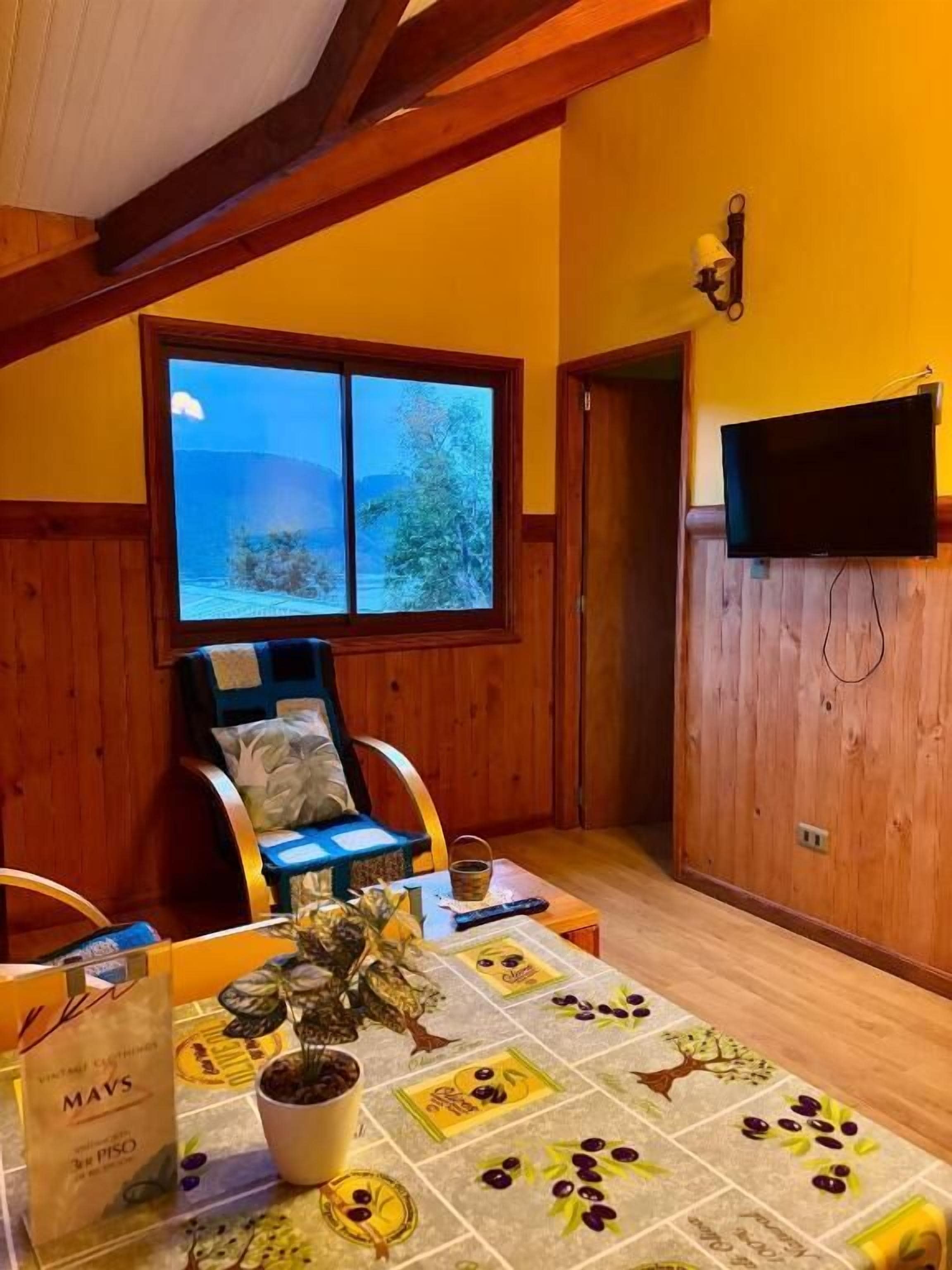 Una habitación con techo de madera, ventana con vistas a montañas, televisor montado en la pared y una mesa con mantel floral.