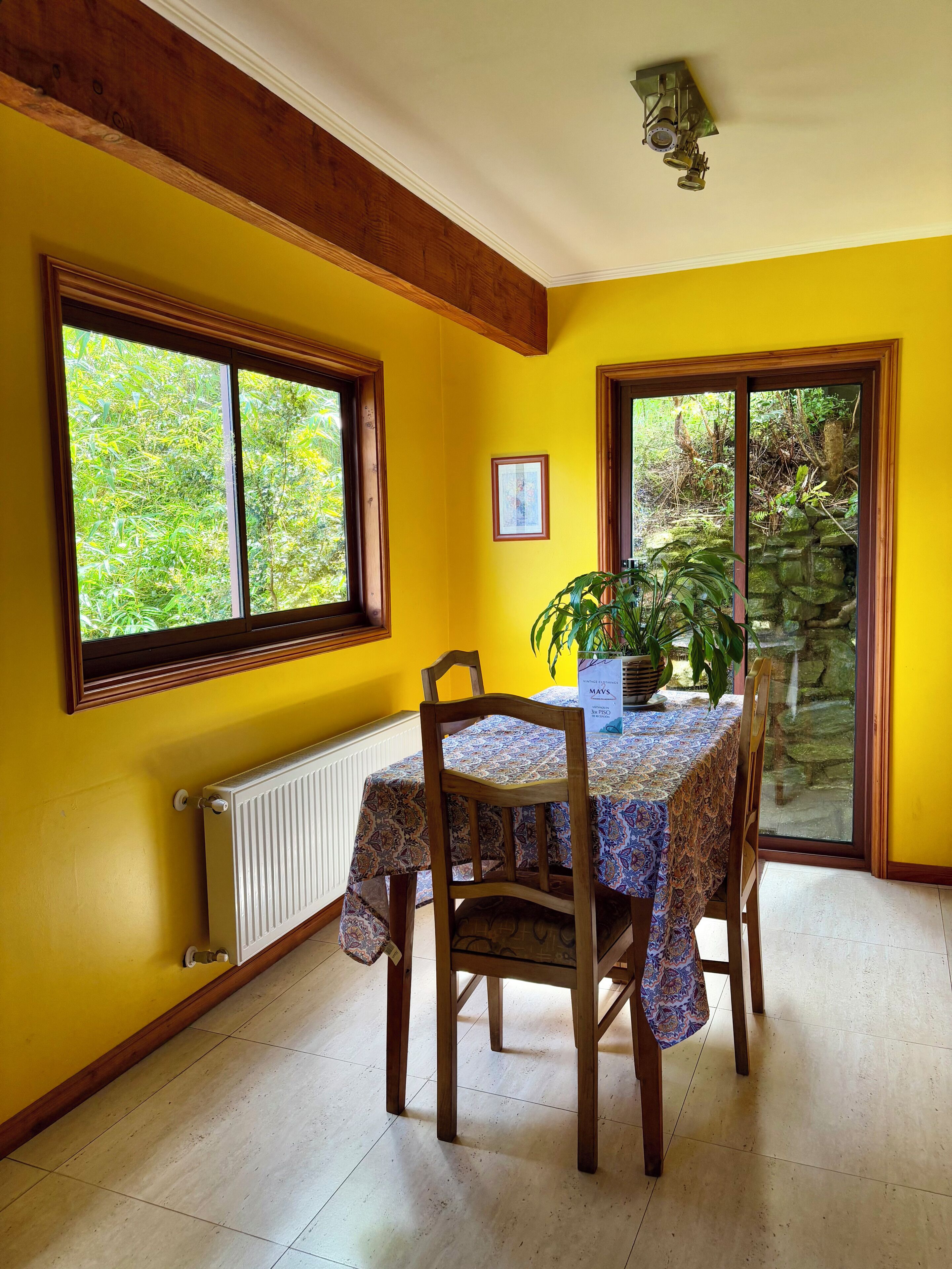 A dining area with a table and chairs, a radiator, and a window with a view of greenery.