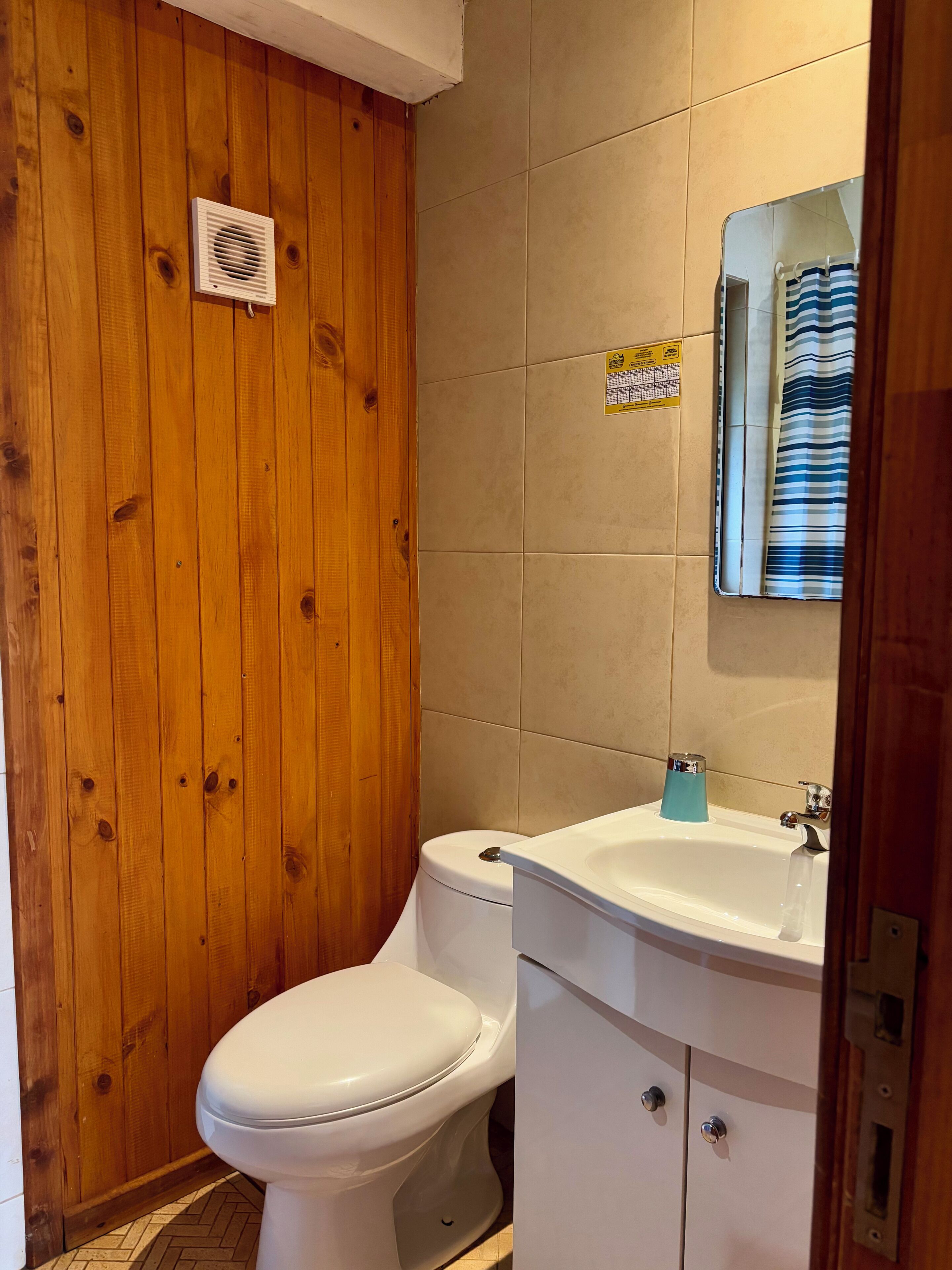 A bathroom with a wooden wall, a white toilet, a sink with a blue soap dispenser, and a mirror.