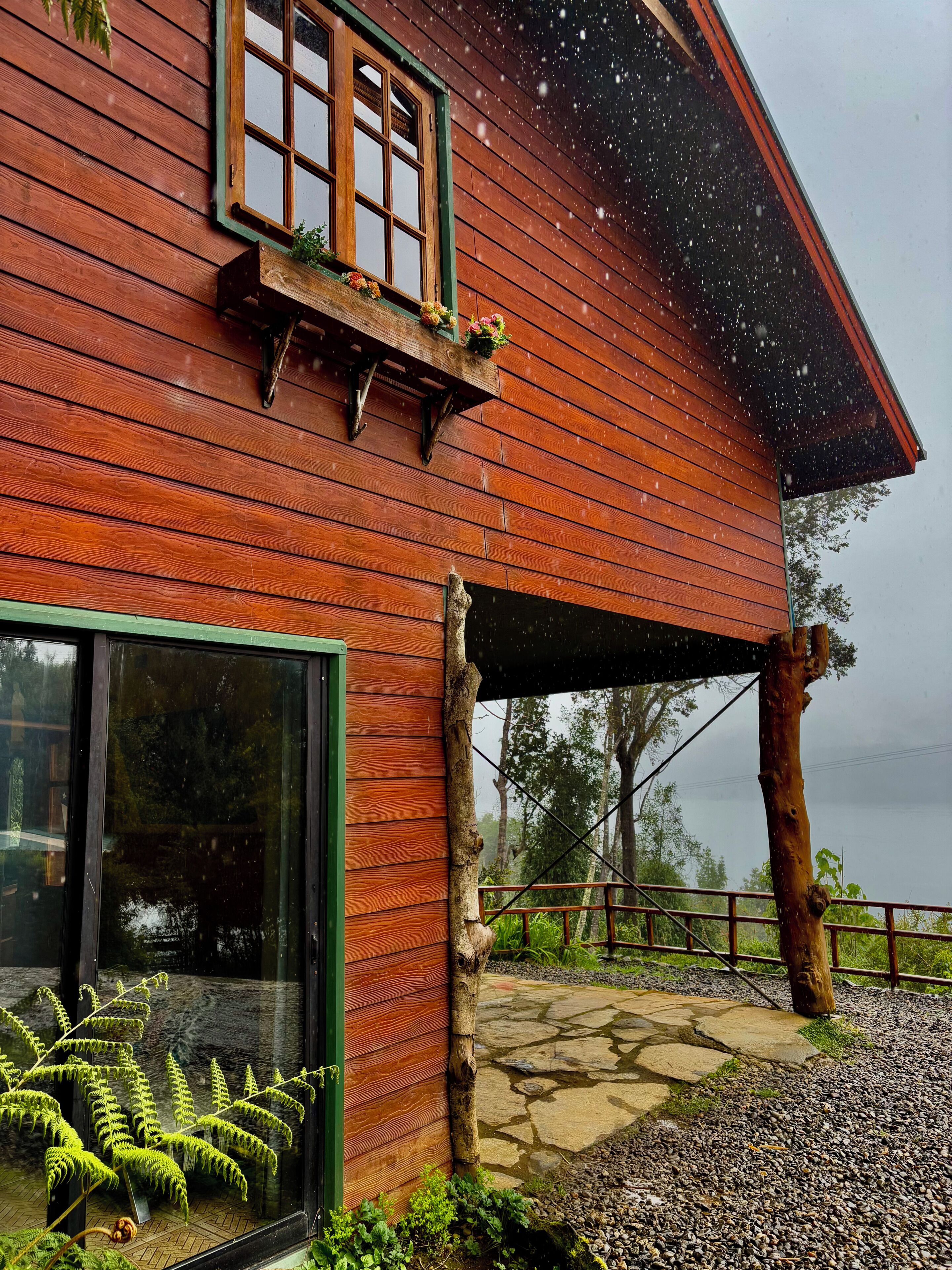 A wooden cabin with a window, a balcony with flowers, and a stone pathway leading to a wooden fence.