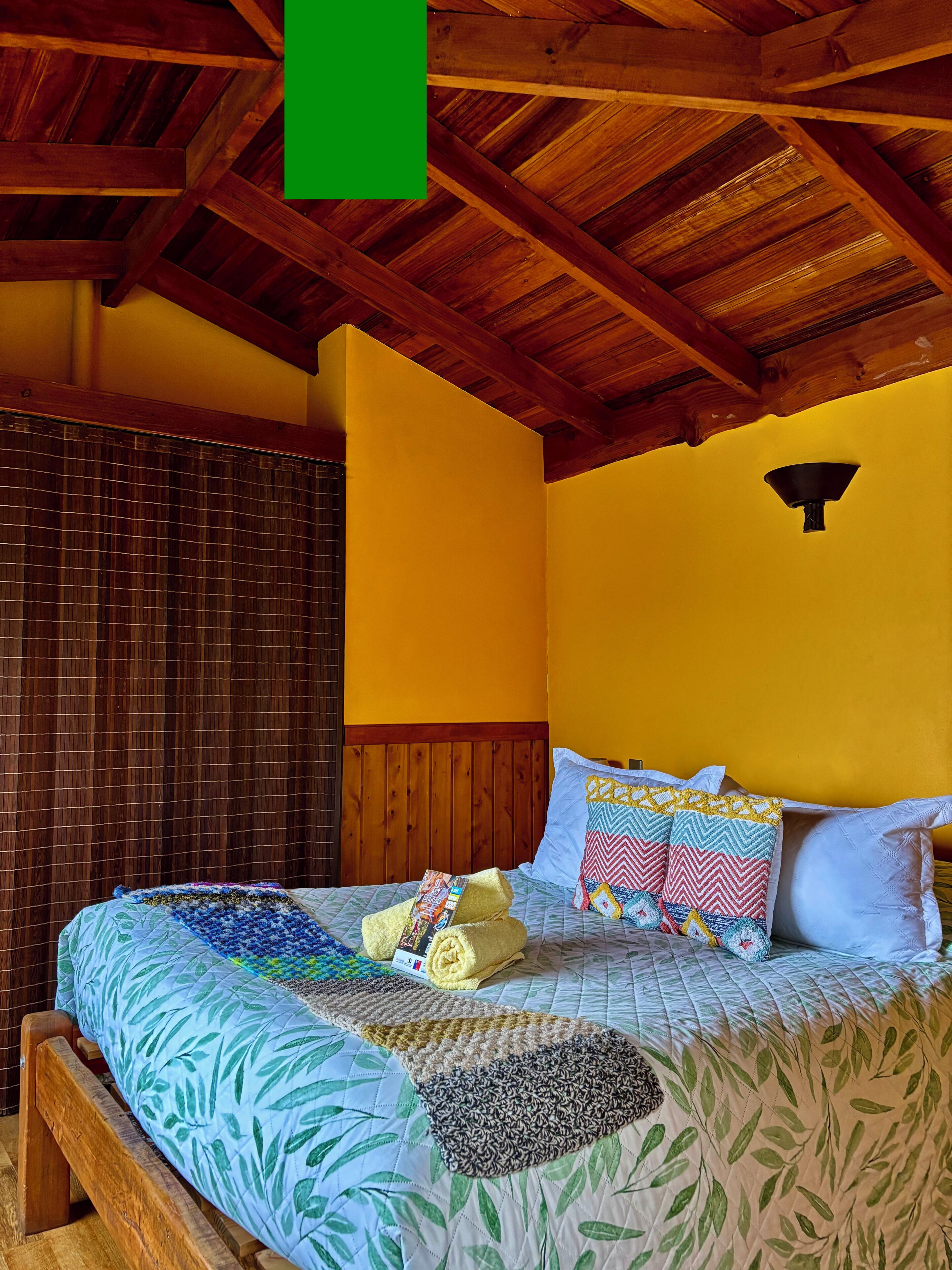 A room with a wooden ceiling, a bed with a quilt, pillows, and a wall-mounted light fixture.