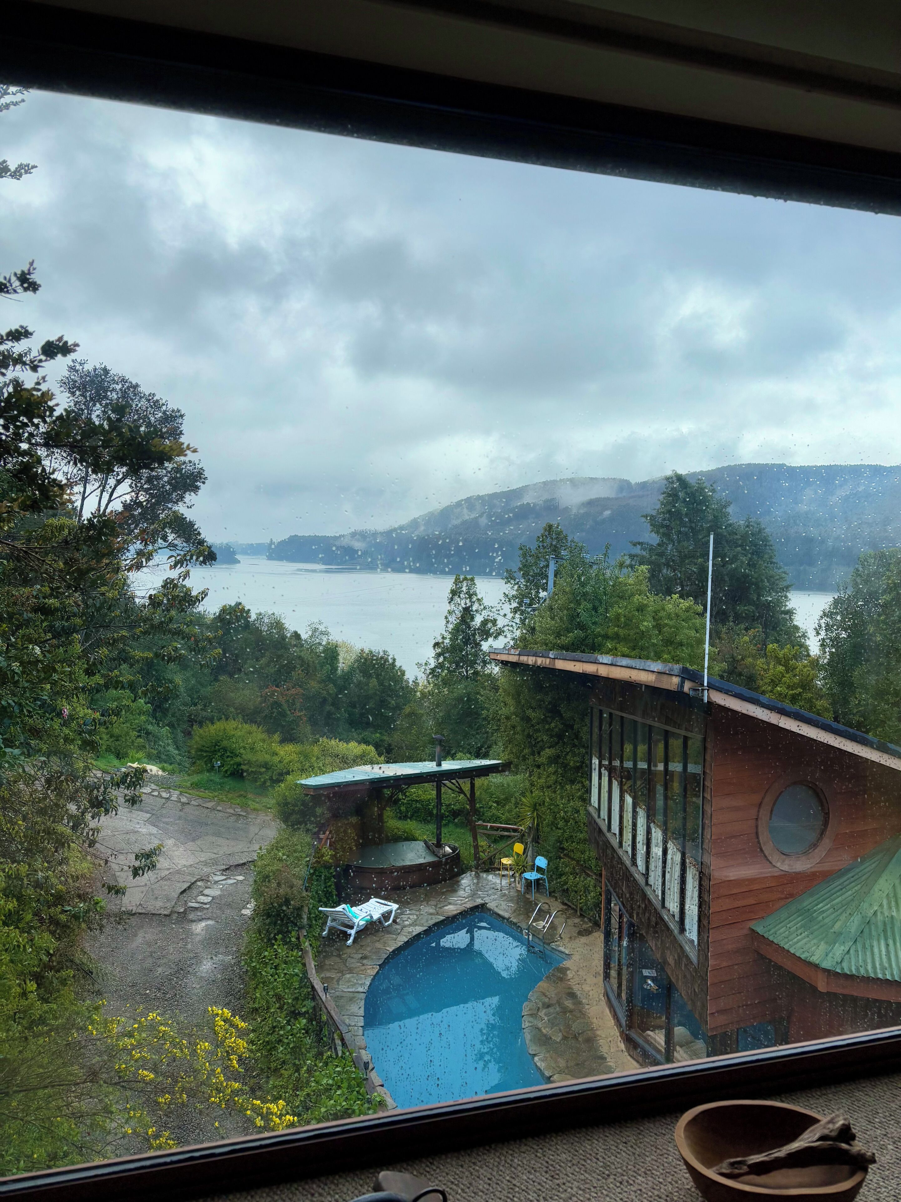 Vista de una zona de piscina con un edificio de madera, un área de descanso techada y vistas al agua y colinas.