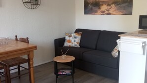 Una sala de estar con una mesa de comedor de madera, un sofá negro, una pequeña mesita auxiliar, un cuadro en la pared y un mueble blanco.