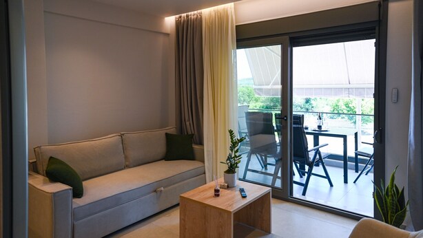 A modern living room with a sofa, a coffee table, and a sliding glass door leading to an outdoor patio. - Estelle luxury living Premium Apartment in Paralia Dionisiou (102) (Paralia Dionisiou)