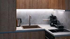 A modern kitchen with wooden cabinets, a stainless steel sink, a coffee maker, and a built-in oven. - Estelle luxury living Premium Apartment in Paralia Dionisiou (102) (Paralia Dionisiou)