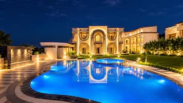 A large, well-lit swimming pool with a reflection of the building in the water, surrounded by a landscaped garden and a grand building with multiple arches and balconies.