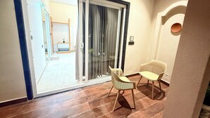 Quarto com porta de vidro deslizante que dá para uma varanda, duas cadeiras, uma mesa pequena e luminária fixada na parede.