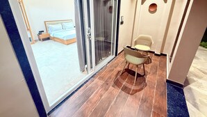 Quarto de hotel com porta de vidro deslizante para uma varanda, cama com cabeceira de madeira, pequena mesa com duas cadeiras e luminária fixa na parede.