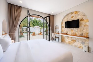 Een slaapkamer met een groot bed, een televisie bevestigd aan een stenen muur, een bureau met een stoel en uitzicht op het buitenleven door een glazen deur.