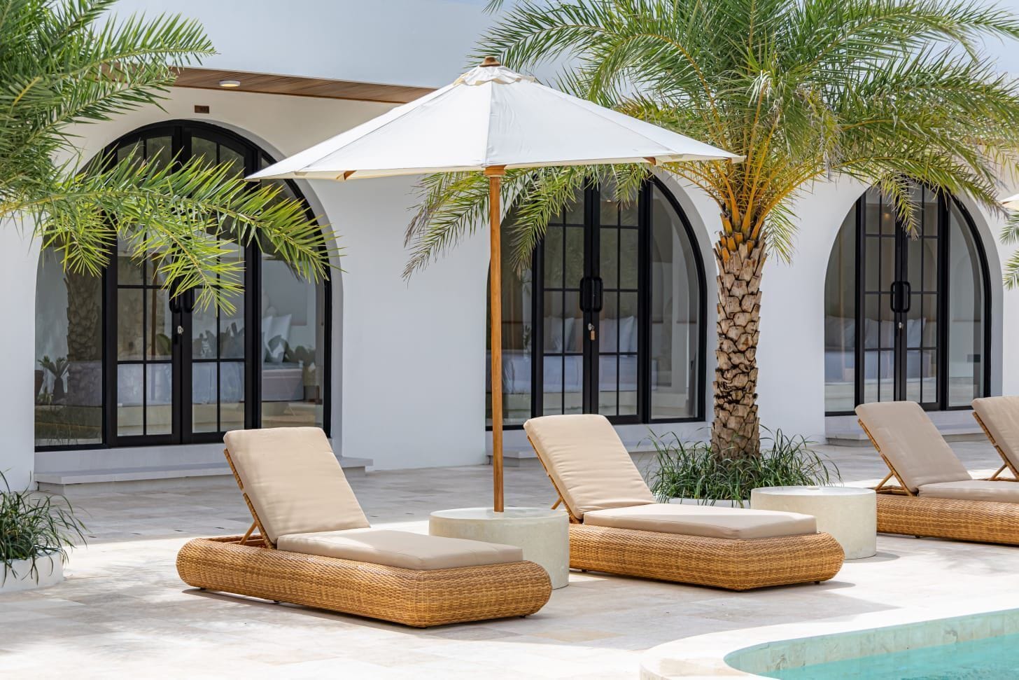 A poolside area with lounge chairs, a white umbrella, and a palm tree.