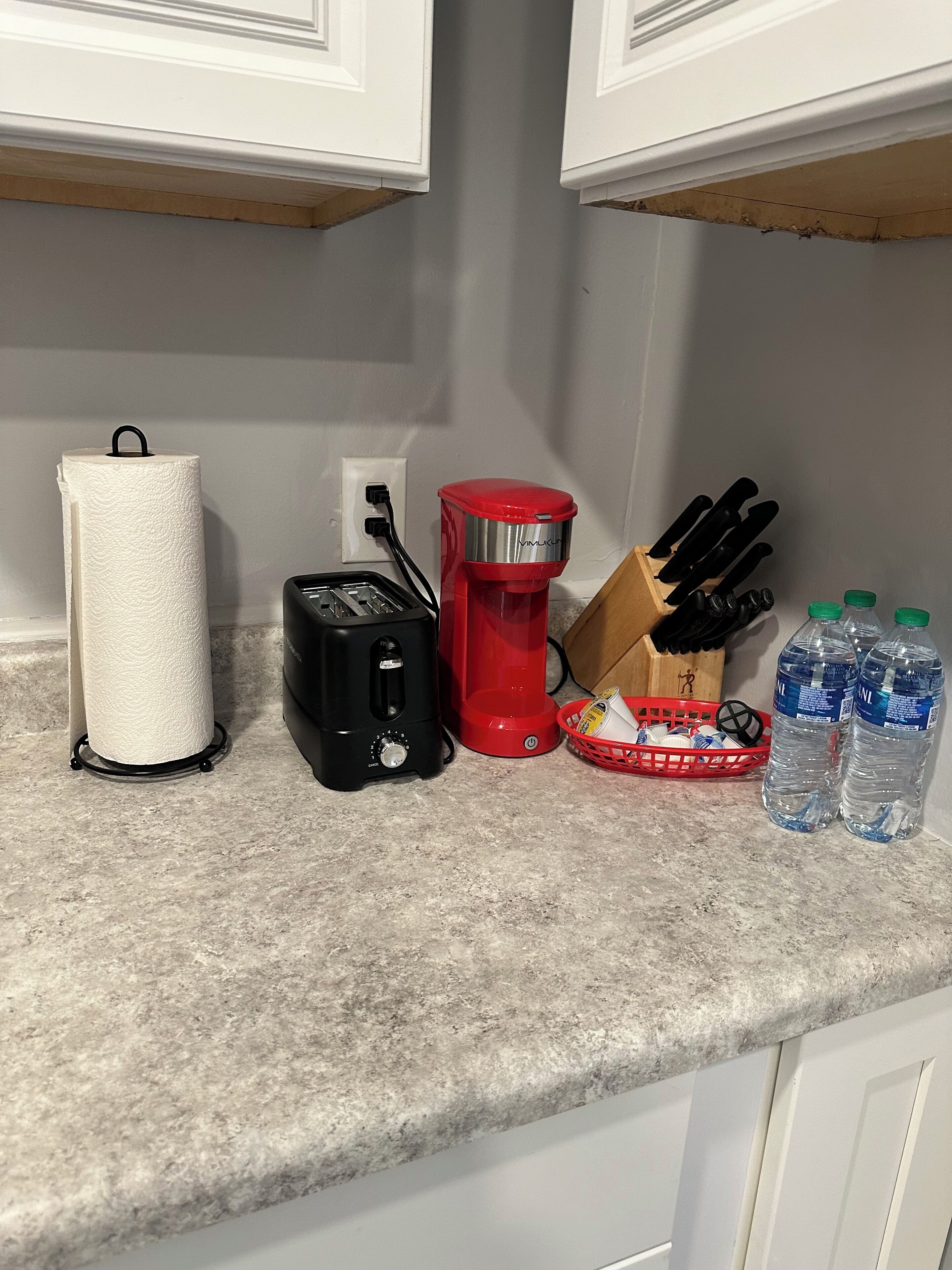 Un plan de travail de cuisine avec un porte-essuie-tout, un grille-pain, une cafetière rouge, un bloc à couteaux et deux bouteilles d’eau.