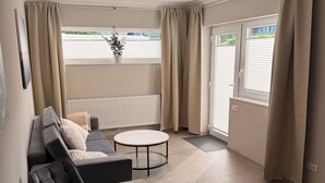 A modern living room with a sofa, a round coffee table, and a dining table. There is a window with curtains, a radiator, and a view of greenery outside.