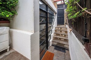 A narrow outdoor corridor with a white wall, a black metal gate, and a staircase leading to a higher level. - Cosy Outhouse in Clapham | Zone 2 | BeeMyGuest® (London)